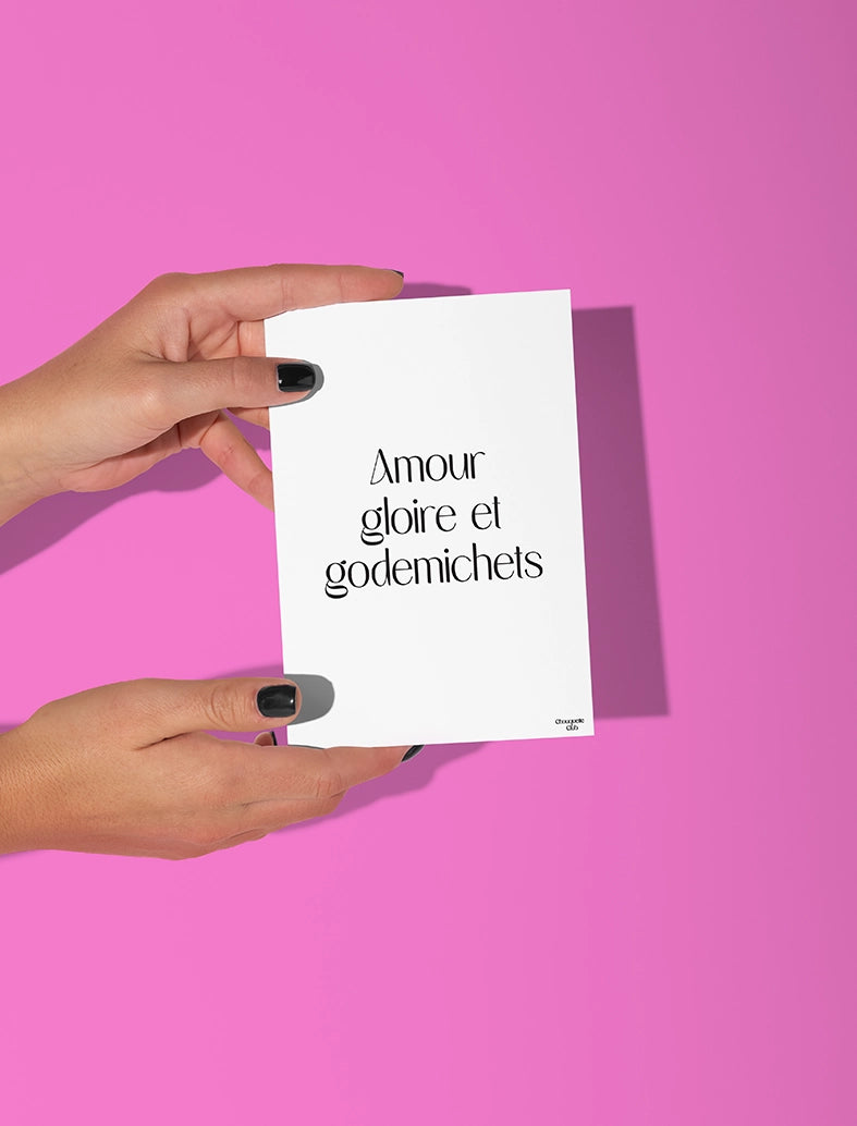 Deux mains qui tiennent une carte postale blanche avec écriture en noir dessus “Amour, gloire et godemichets”