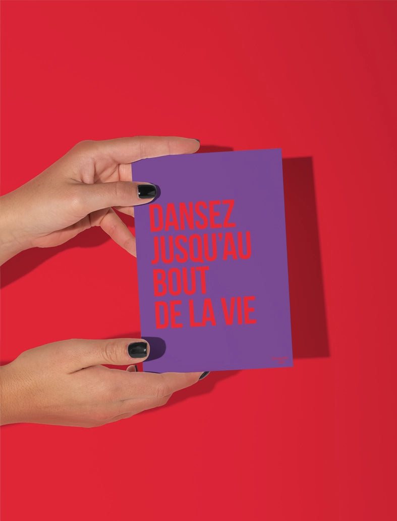 Carte postale écrit "dansez jusqu'au bout de la vie" en rouge sur fond violet tenue par deux mains