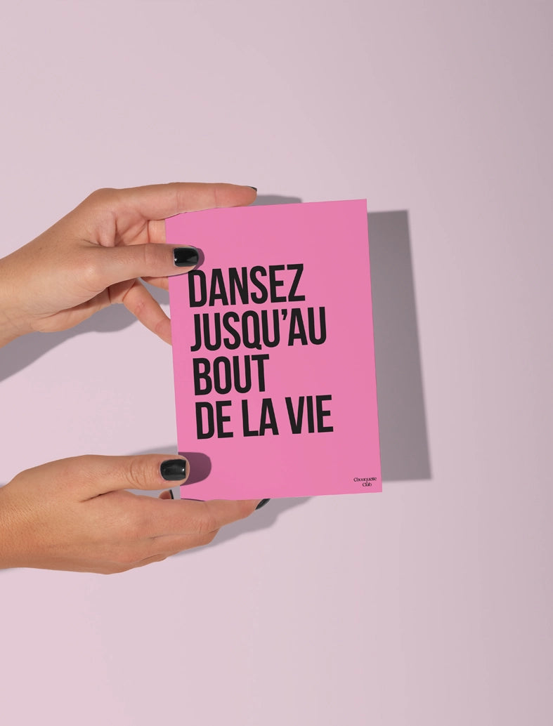 Carte postale écrit "dansez jusqu'au bout de la vie" en noir sur fond rose tenue par deux mains
