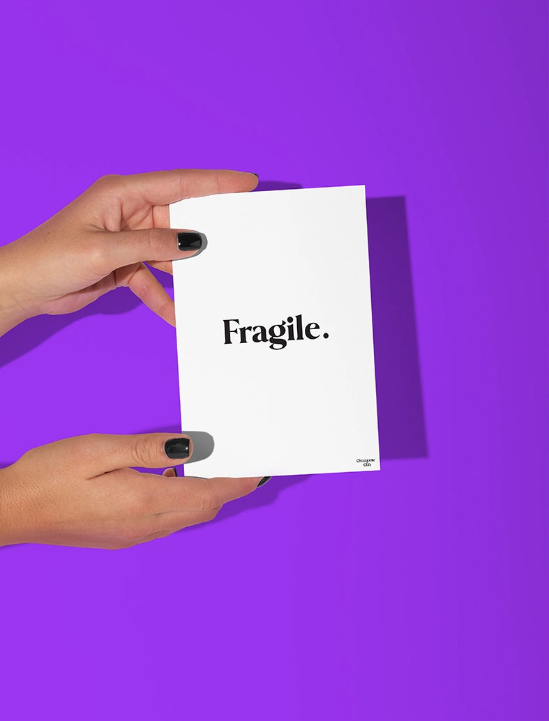 Deux mains qui tiennent une carte postale blanche avec écriture en noir dessus “Fragile”