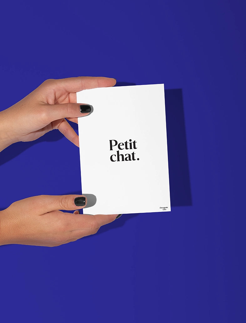 Deux mains qui tiennent une carte postale blanche avec écriture en noir dessus “Petit chat”