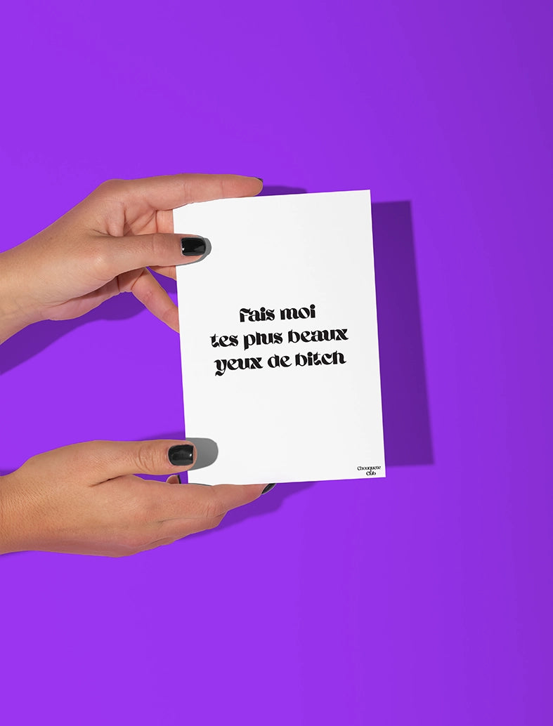 Deux mains qui tiennent une carte postale blanche avec écriture en noir dessus “Fais moi tes plus beaux yeux de bitch”