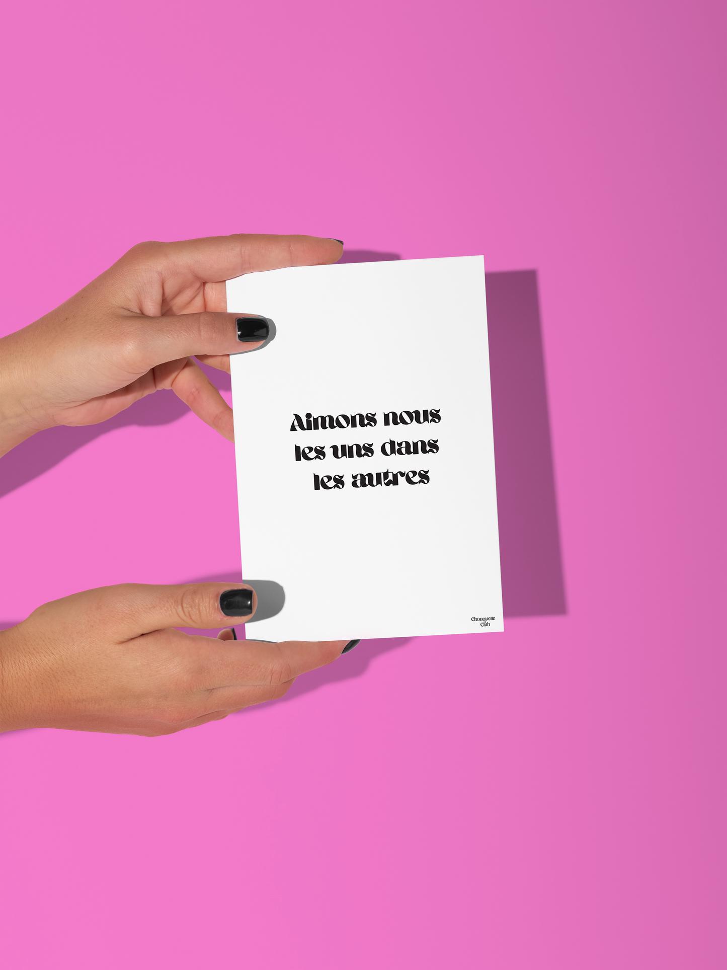 Deux mains qui tiennent une carte postale blanche avec écriture en noir dessus “Aimons nous les uns dans les autres”.