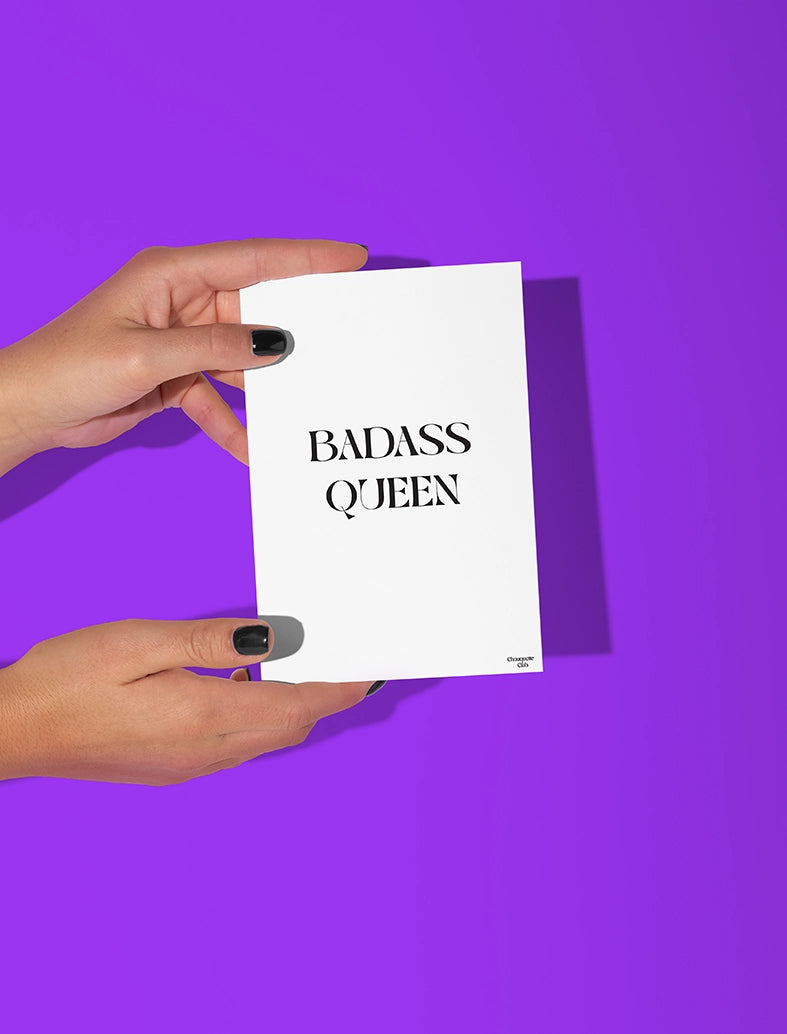 Deux mains qui tiennent une carte postale blanche avec écriture en noir dessus “Badass Queen”