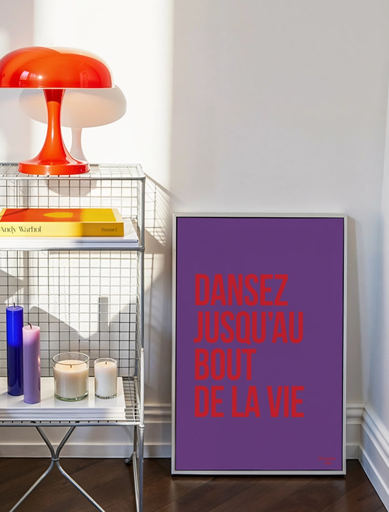 Dansez jusqu'au bout de la vie affiche (violet et rouge) posé au sol à coté d'une lampe orange posée sur une étagère métallique