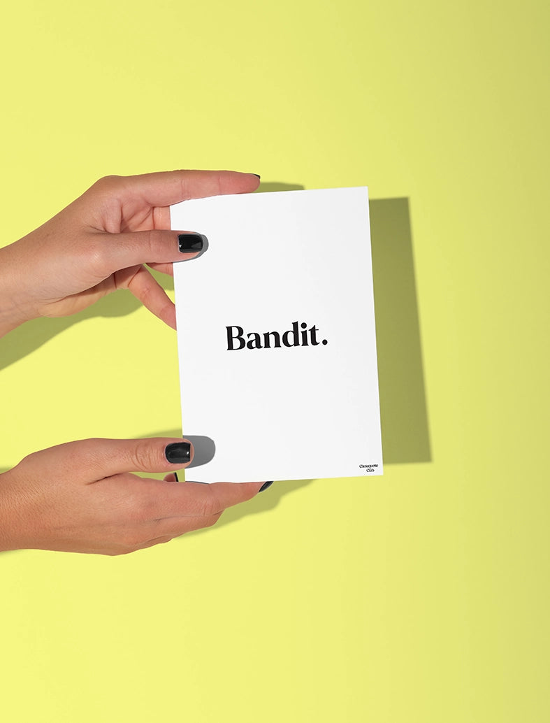 Deux mains qui tiennent une carte postale blanche avec écriture en noir dessus “Bandit ”
