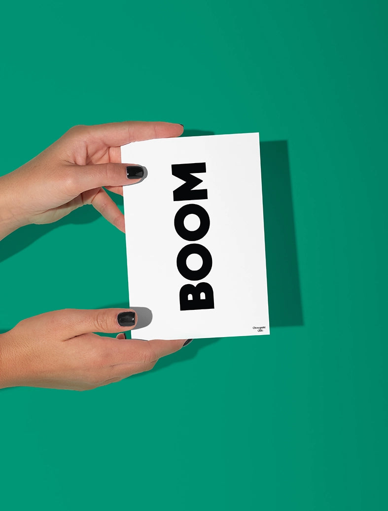 Deux mains qui tiennent une carte postale blanche avec écriture en noir dessus “Boom”.