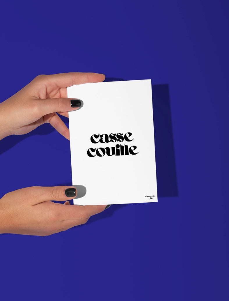 Deux mains qui tiennent une carte postale blanche avec écriture en noir dessus "casse couille"
