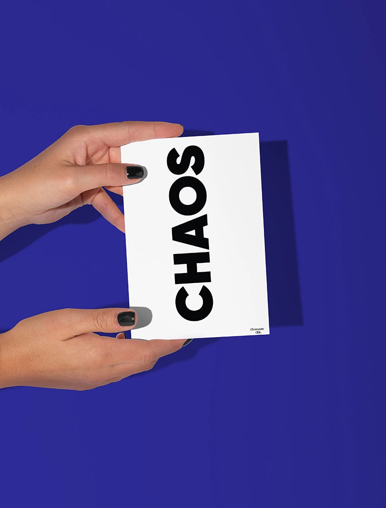 Deux mains qui tiennent une carte postale blanche avec écriture en noir dessus “Chaos”