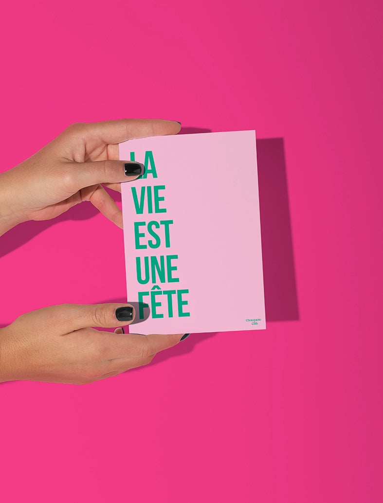 Carte postale rose tenue par deux mains, avec le texte vert '"la vie est une fête"