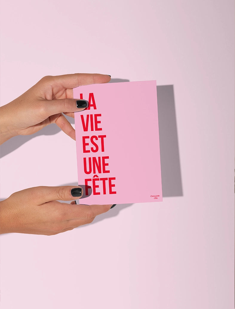 Carte postale rose tenue par deux mains, avec le texte rouge '"la vie est une fête"