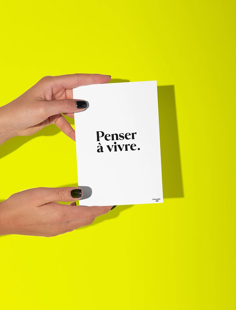 Deux mains qui tiennent une carte postale blanche avec écriture en noir dessus “ Penser à vivre”