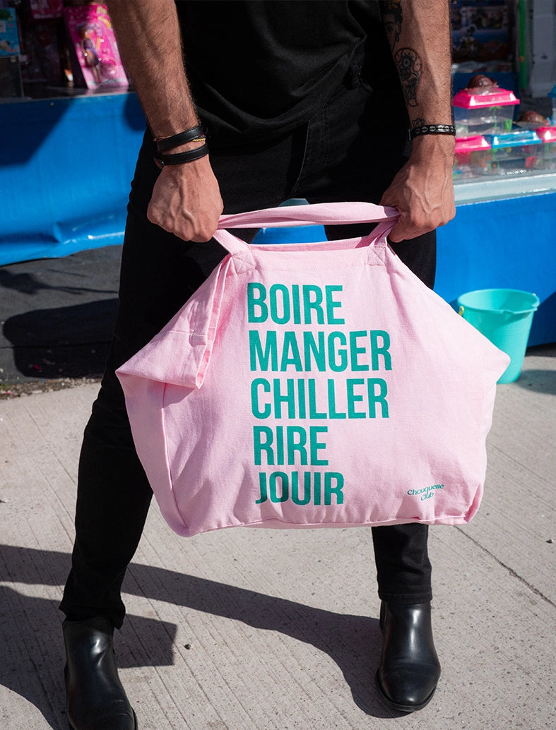 Le modéle homme brun porte un gros sac en coton rose écrit dessus "boire manger chiller rire jouir" en vert de chez Chouquette Club