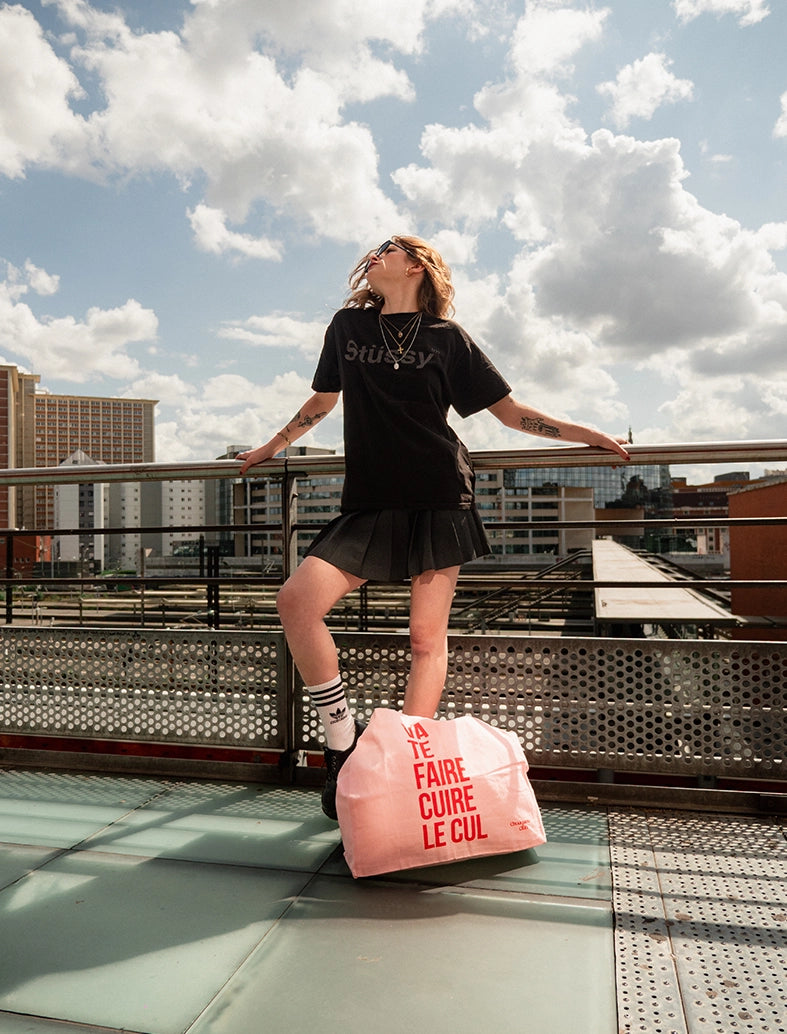 sac en coton rose et rouge de chouquette club marqué dessus "va te faire cuire le cul" porté par une fille brune aux longs cheveux