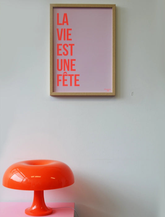 La vie est une fête orange fluo et rose pâle affiche A3 accroché au mur blanc posé sur un meuble rose avec lampe orange