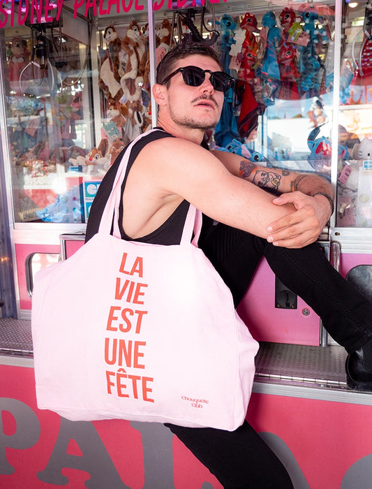 sac en coton rose et rouge de chouquette club marqué dessus "la vie est une fête" porté par une homme dans une fête foraine