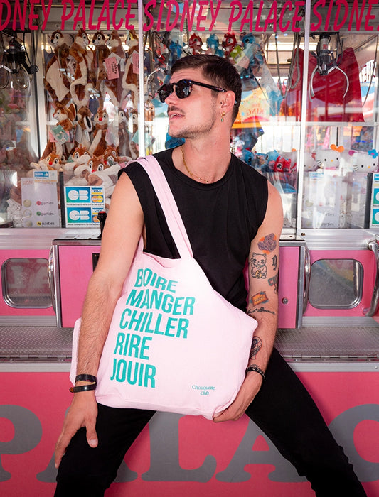 Le modéle homme brun porte un gros sac en coton rose écrit dessus "boire manger chiller rire jouir" en vert de chez Chouquette Club